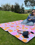 Woman sitting on corner of the retro floral picnic rug, reading a book in grassy park setting