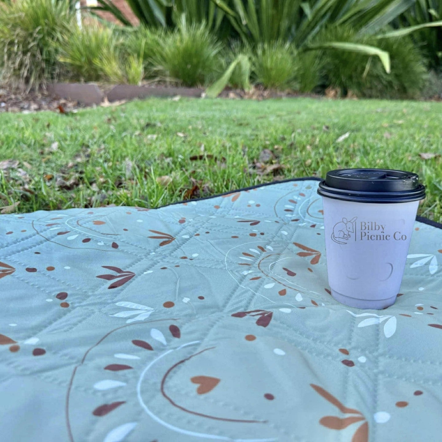 close up of a boho picnic rug corner with a coffee cup resting on it, on grass-Bilby Picnic Co