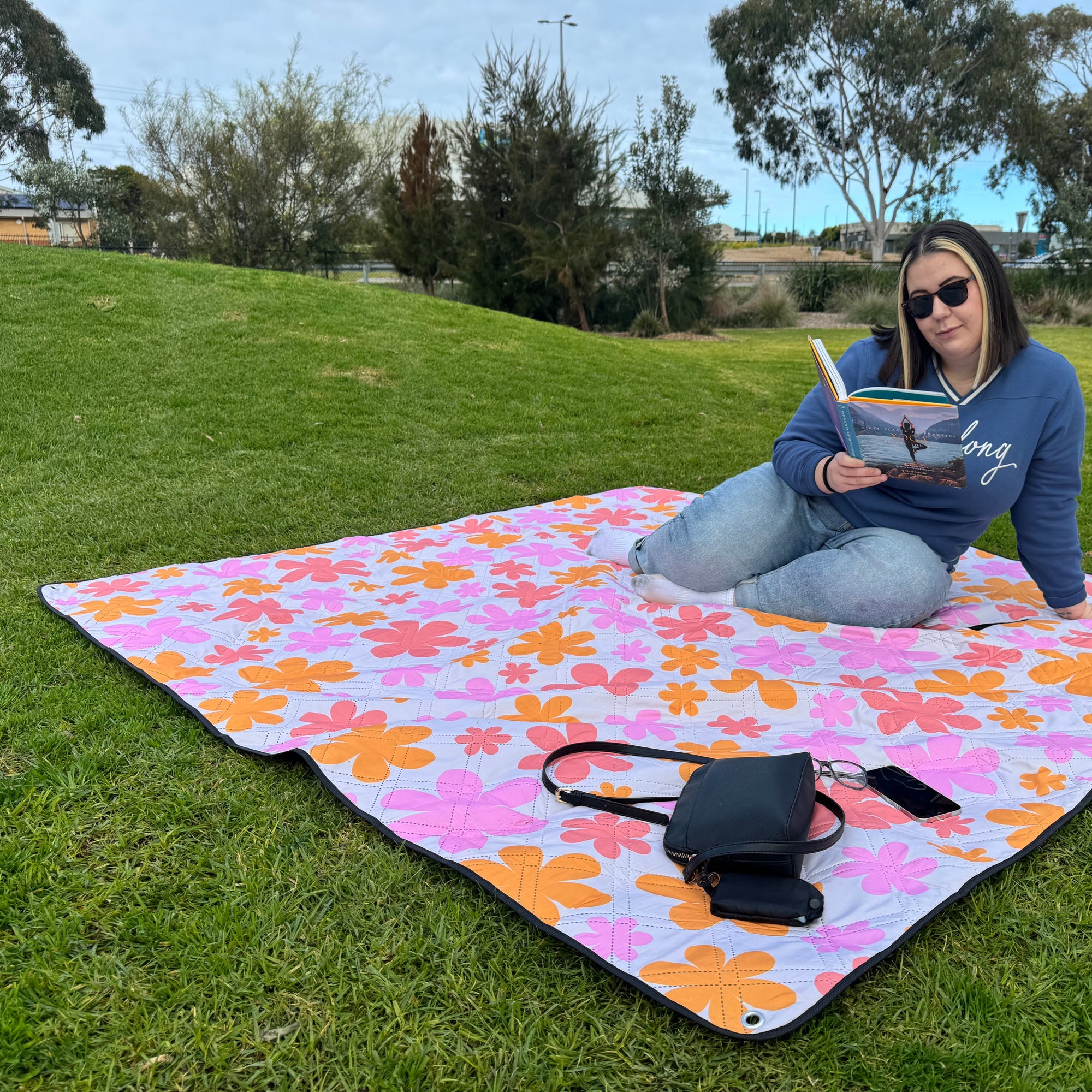 Woman sitting on corner of the retro floral picnic rug, reading a book in grassy park setting